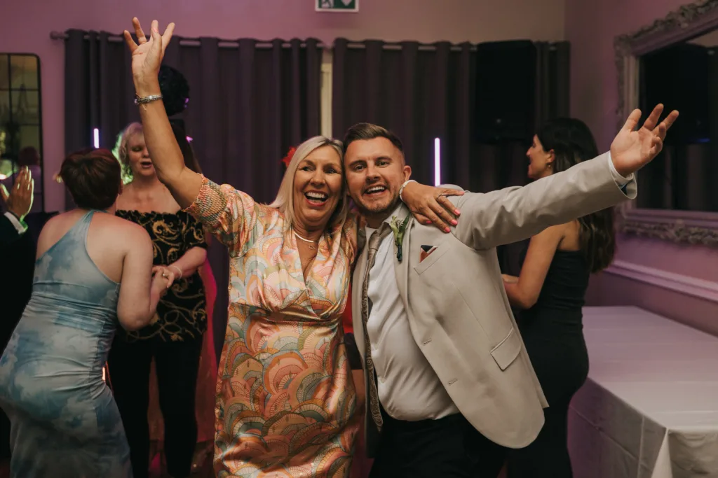 Two people, a woman in a colorful dress and a man in a light suit, stand at the center of a lively Dower House Hotel wedding with arms around each other, smiling and cheering as guests dance in the festive, joyful atmosphere. © Aimee Lince Photography - Wedding photographer in Lincolnshire, Yorkshire & Nottinghamshire