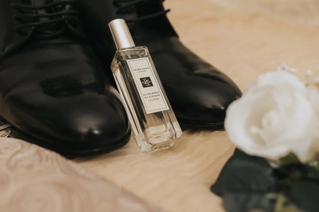 A clear bottle of Jo Malone cologne sits before polished black dress shoes on a beige surface. To the right, a white artificial rose adds elegance—an ideal detail for a wedding at Dower House Hotel captured by a Lincolnshire wedding photographer. © Aimee Lince Photography - Wedding photographer in Lincolnshire, Yorkshire & Nottinghamshire