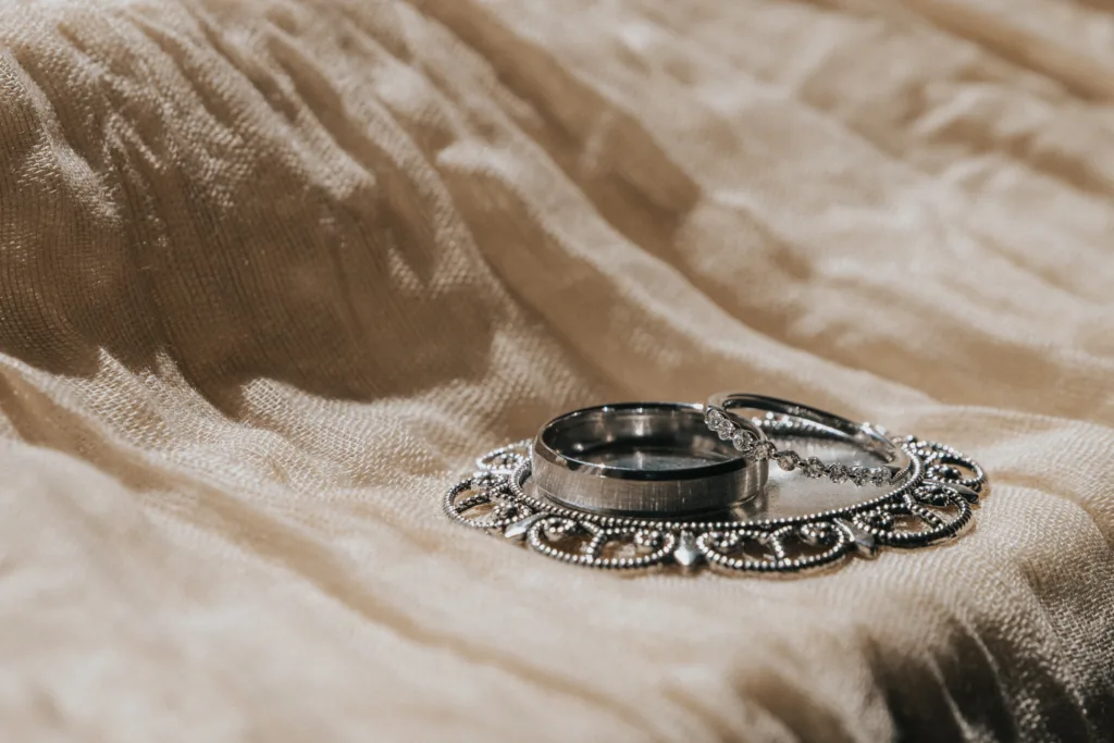 Two silver rings, one plain and one with small stones, rest on an ornate silver pendant atop softly textured beige fabric. Bathed in natural light, this elegant scene captures the romance of a Dower House Hotel wedding in Woodhall Spa. © Aimee Lince Photography - Wedding photographer in Lincolnshire, Yorkshire & Nottinghamshire