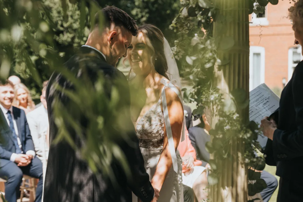 A bride and groom stand facing each other, smiling and holding hands during a Dower House Hotel wedding ceremony. Sunlight filters through greenery as guests watch in the background and an officiant holds papers on the right. © Aimee Lince Photography - Wedding photographer in Lincolnshire, Yorkshire & Nottinghamshire