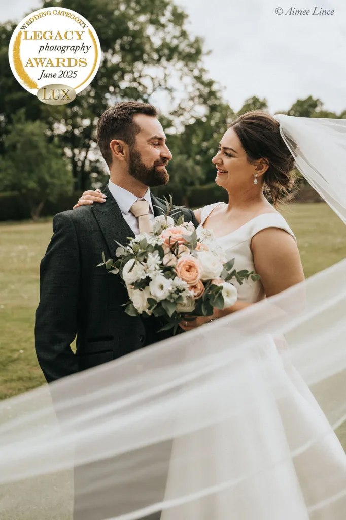 A bride in a white dress and veil smiles at her groom as they embrace outdoors. She holds a pastel bouquet. Both look happy. A gold “Legacy Photography Awards June 2025” badge highlights this UK wedding photographer’s storytelling wedding photography. © Aimee Lince Photography - Wedding photographer in Lincolnshire, Yorkshire & Nottinghamshire