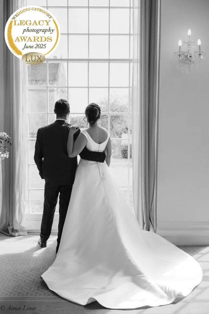 A bride and groom stand side by side, backs to the camera, gazing out a tall window bathed in soft light. This storytelling wedding photography scene earned an award badge for “Legacy Photography Awards June 2025 LUX” at the top left. © Aimee Lince Photography - Wedding photographer in Lincolnshire, Yorkshire & Nottinghamshire