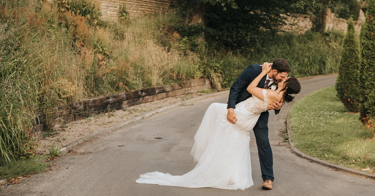 A groom in a navy suit dips and kisses his bride in a white lace dress on a paved path at Hackness Grange. Lush greenery, tall grass, and a stone wall line the background, capturing this romantic Scarborough Wedding moment. © Aimee Lince Photography - Wedding photographer in Lincolnshire, Yorkshire & Nottinghamshire