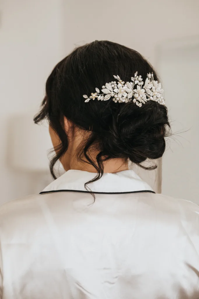 A woman with dark hair styled in a low, loose bun, adorned with a decorative silver floral hairpiece, stands in a softly lit room. Captured by a Scarborough wedding photographer at Hackness Grange, she wears a silky white robe with black piping. © Aimee Lince Photography - Wedding photographer in Lincolnshire, Yorkshire & Nottinghamshire