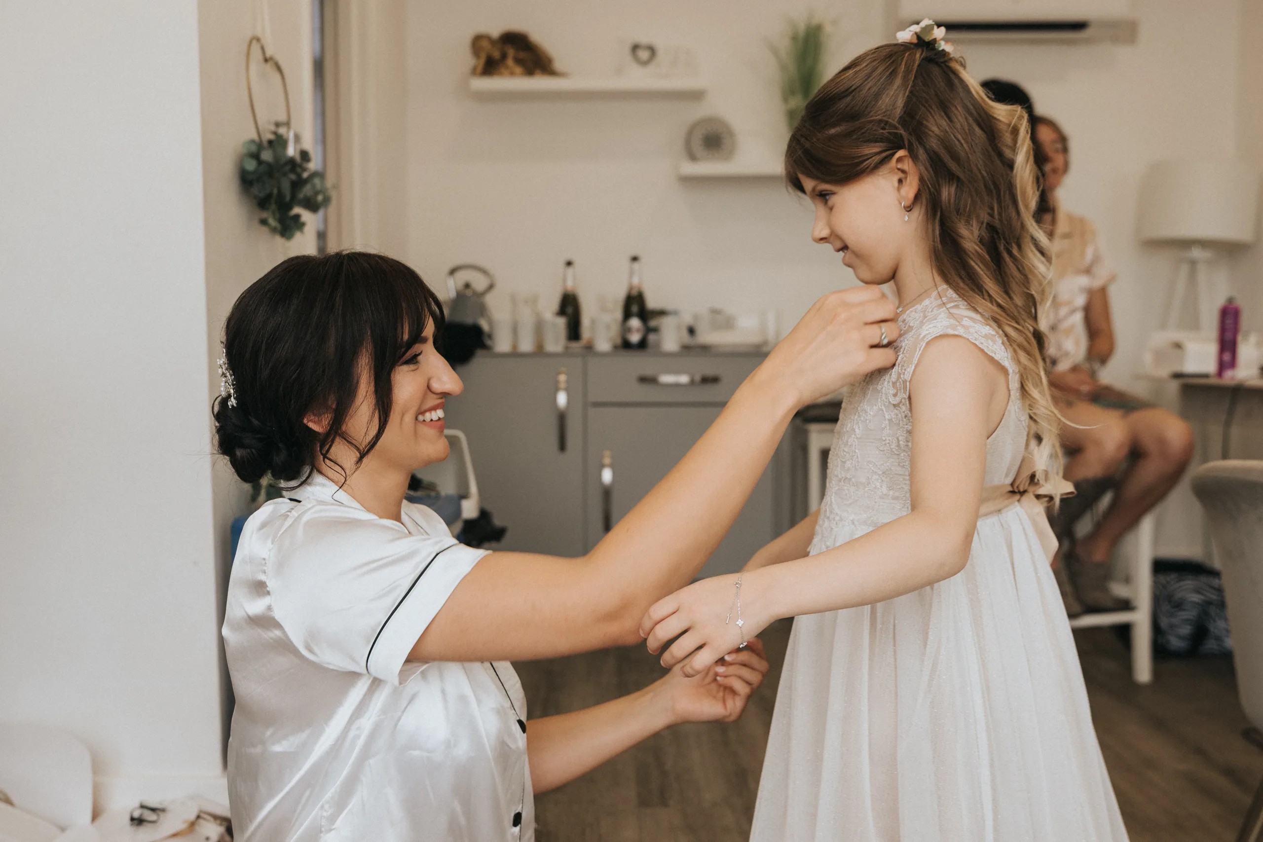 shane-carina-wedding-photography-scarborough-hackness-grange-photographer-copyright-aimee-lince-106-of-649-©-aimee-lince-photography-16-07-2025 A woman in a satin robe kneels, smiling as she helps a young girl in an ivory dress adjust her necklace. Captured by a Scarborough wedding photographer, the sweet moment unfolds in a light, modern room at Hackness Grange. © Aimee Lince Photography - Wedding photographer in Lincolnshire, Yorkshire & Nottinghamshire