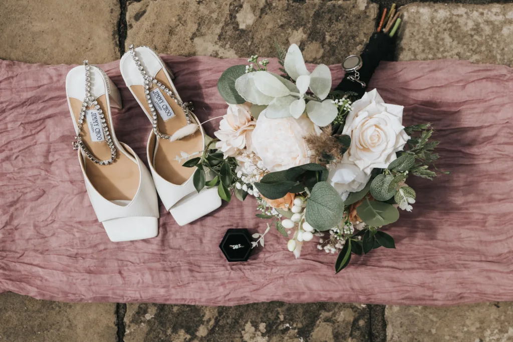 A pair of white, square-toe shoes with jeweled straps sits on a mauve cloth beside a bouquet of white and blush flowers, greenery, and a black ring box—beautifully styled by a Hackness Grange wedding photographer on stone pavement. © Aimee Lince Photography - Wedding photographer in Lincolnshire, Yorkshire & Nottinghamshire