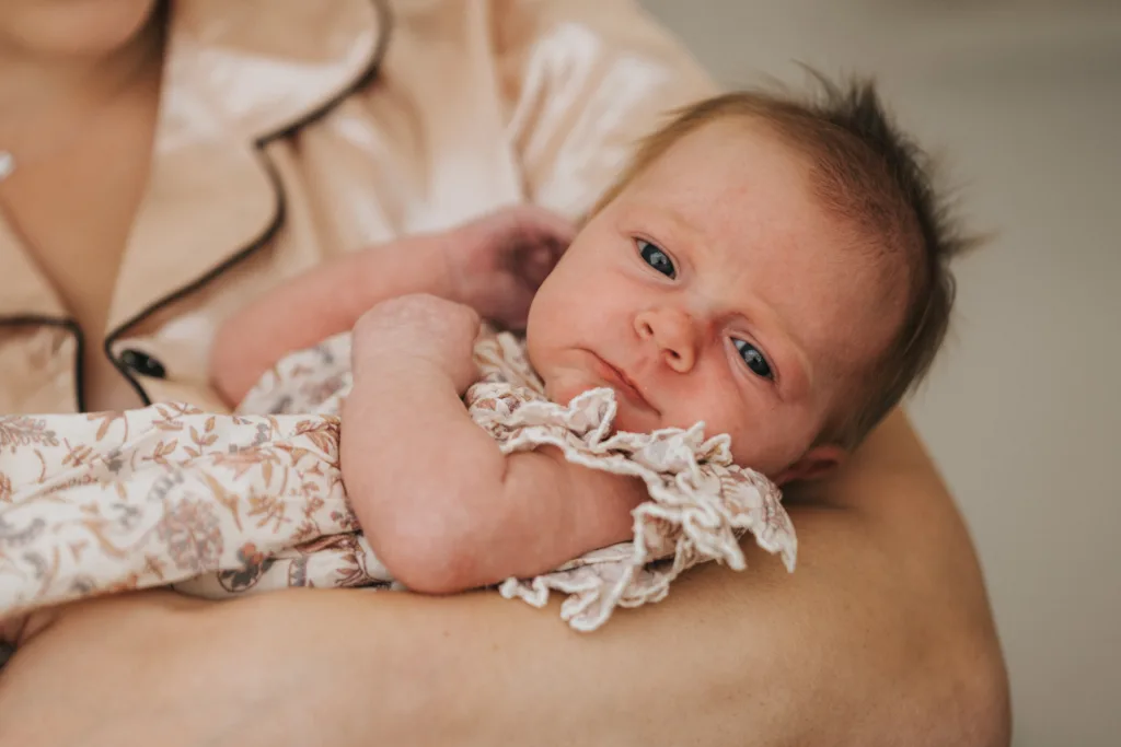 A newborn baby with blue eyes and light brown hair is cradled in an adult's arms. Captured by a wedding photographer in Scarborough, the baby wears a frilly, floral-patterned outfit and gazes calmly at the camera against a soft, neutral background. © Aimee Lince Photography - Wedding photographer in Lincolnshire, Yorkshire & Nottinghamshire