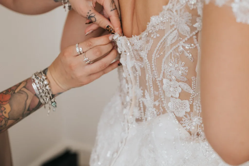 Close-up of two hands fastening delicate lace buttons on the back of a bride’s intricately embroidered dress. Captured by a wedding photographer at Hackness Grange near Scarborough, tattoos and jewelry add personality to the touching moment. © Aimee Lince Photography - Wedding photographer in Lincolnshire, Yorkshire & Nottinghamshire