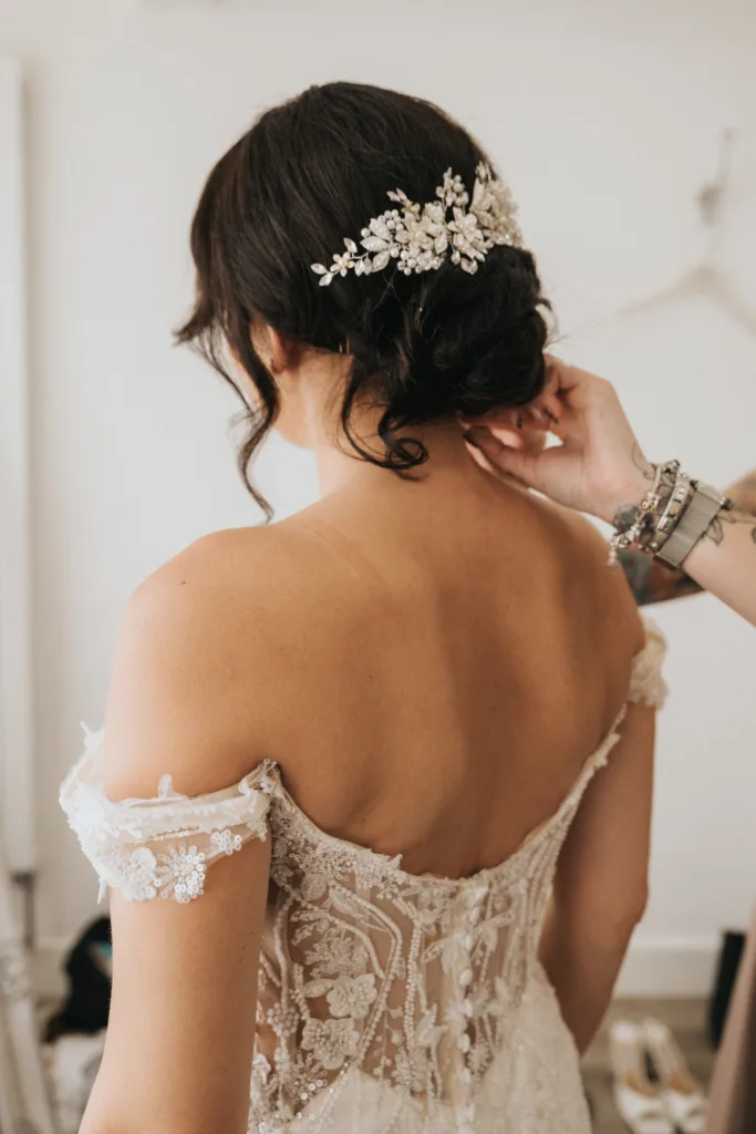 A bride at Hackness Grange, seen from behind, wears an off-the-shoulder lace gown. Her dark hair is in an elegant updo with a sparkly floral hairpiece as a wedding photographer in Scarborough captures someone adjusting her dress strap. The background is softly blurred. © Aimee Lince Photography - Wedding photographer in Lincolnshire, Yorkshire & Nottinghamshire