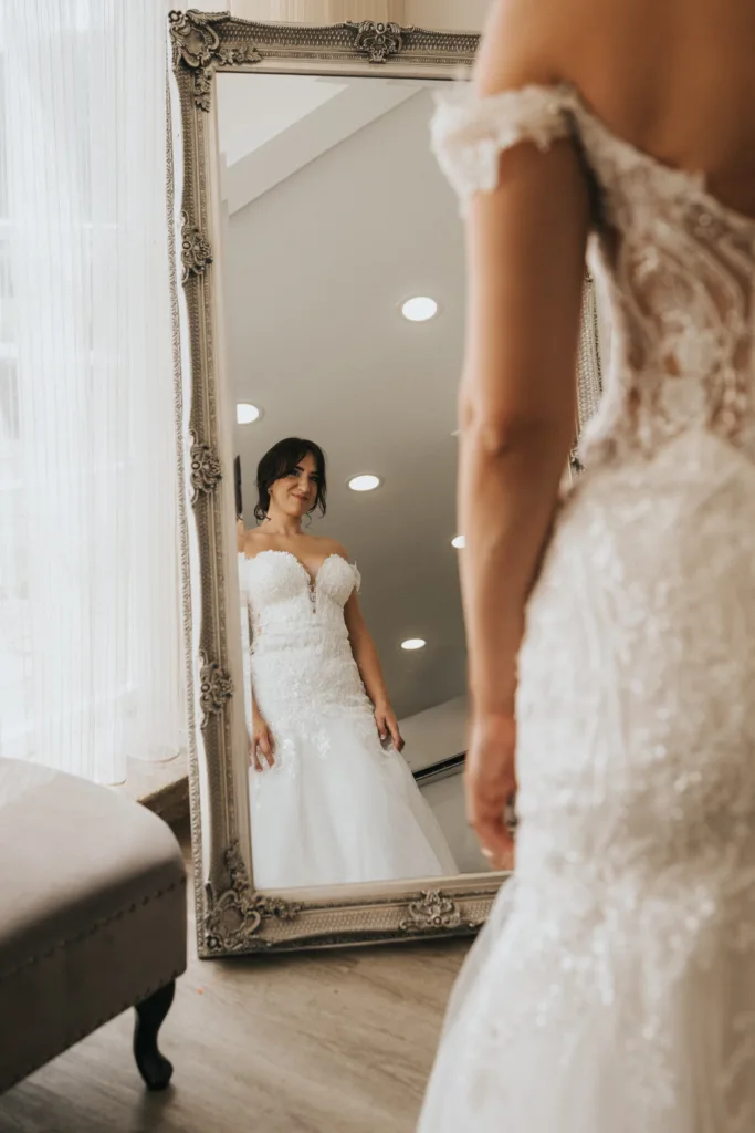 A bride in an off-the-shoulder, lace wedding gown smiles at her reflection in a large ornate mirror at Hackness Grange near Scarborough. The room is softly lit, and a velvet bench sits beside her—perfect for a wedding photographer to capture the moment. © Aimee Lince Photography - Wedding photographer in Lincolnshire, Yorkshire & Nottinghamshire