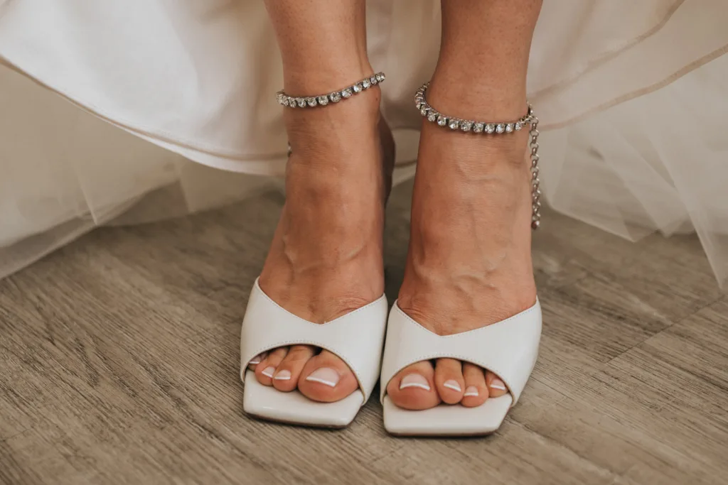 A close-up of a woman's feet wearing white, open-toe heels and sparkly anklets. Perfect for a wedding photographer in Scarborough, her neatly painted toenails peek out on the wooden floor beneath the hem of a white dress. © Aimee Lince Photography - Wedding photographer in Lincolnshire, Yorkshire & Nottinghamshire