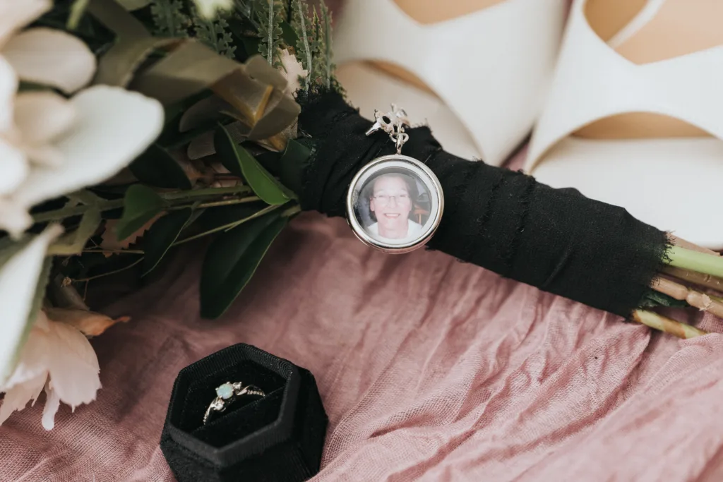 A bridal bouquet wrapped in black ribbon, perfect for a Hackness Grange celebration near Scarborough, holds a round photo charm of a smiling elderly woman. Next to it sits an open black ring box with a silver engagement ring on pink textured fabric. © Aimee Lince Photography - Wedding photographer in Lincolnshire, Yorkshire & Nottinghamshire