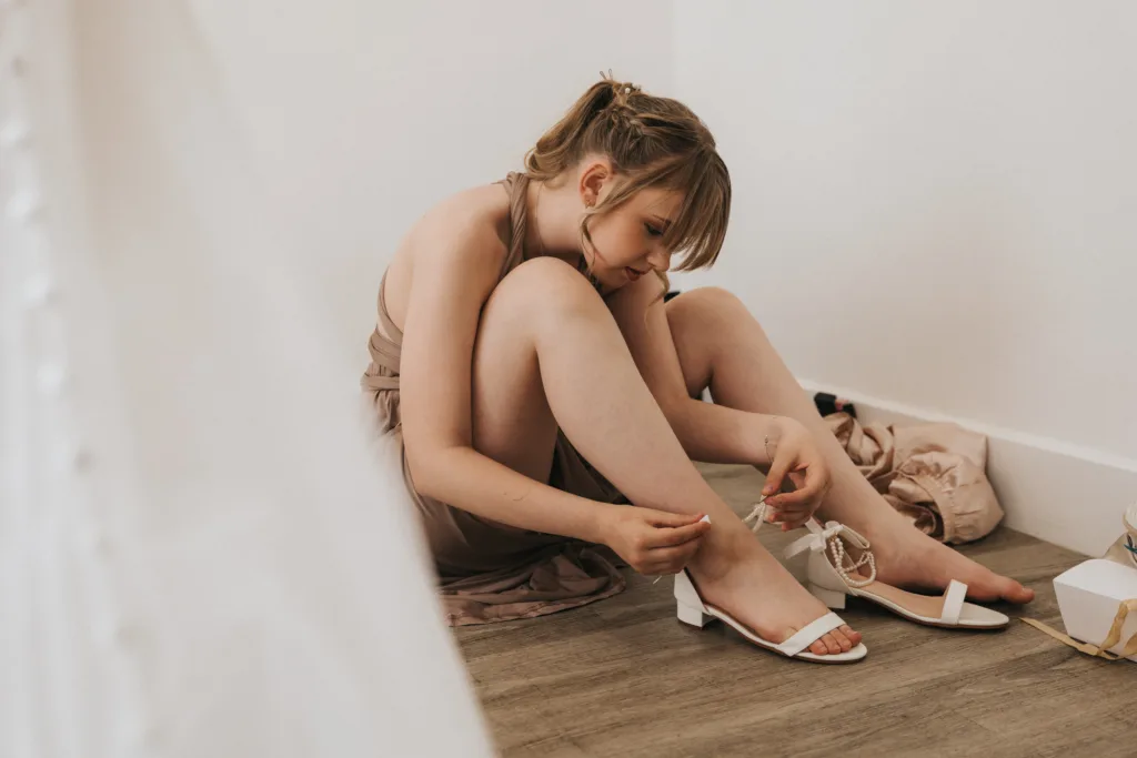 A young woman in a taupe dress sits on a wooden floor at Hackness Grange, leaning against a white wall, putting on white heeled sandals. Clothes and a shoebox are scattered nearby. Her light brown hair is tied back as she focuses on fastening her shoe. © Aimee Lince Photography - Wedding photographer in Lincolnshire, Yorkshire & Nottinghamshire