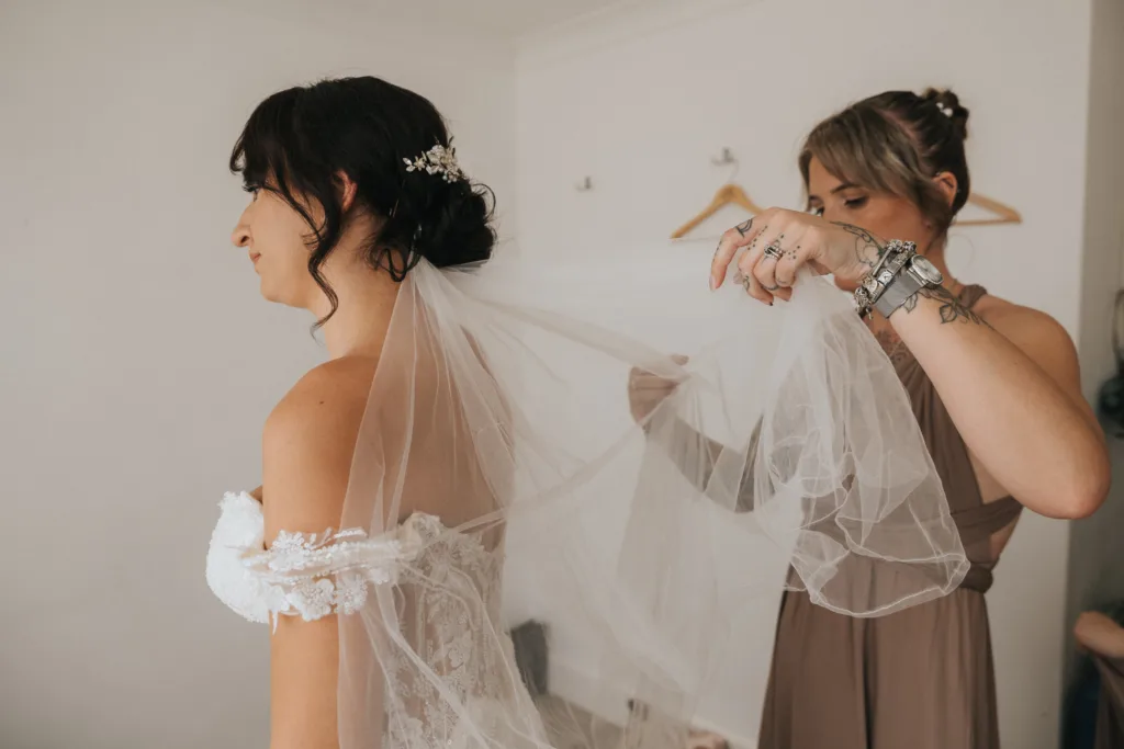 A bride in an off-shoulder lace wedding dress stands as another woman adjusts her long, sheer veil. Captured by a wedding photographer at Hackness Grange, the scene features simple white walls and two empty wooden hangers in the background. © Aimee Lince Photography - Wedding photographer in Lincolnshire, Yorkshire & Nottinghamshire