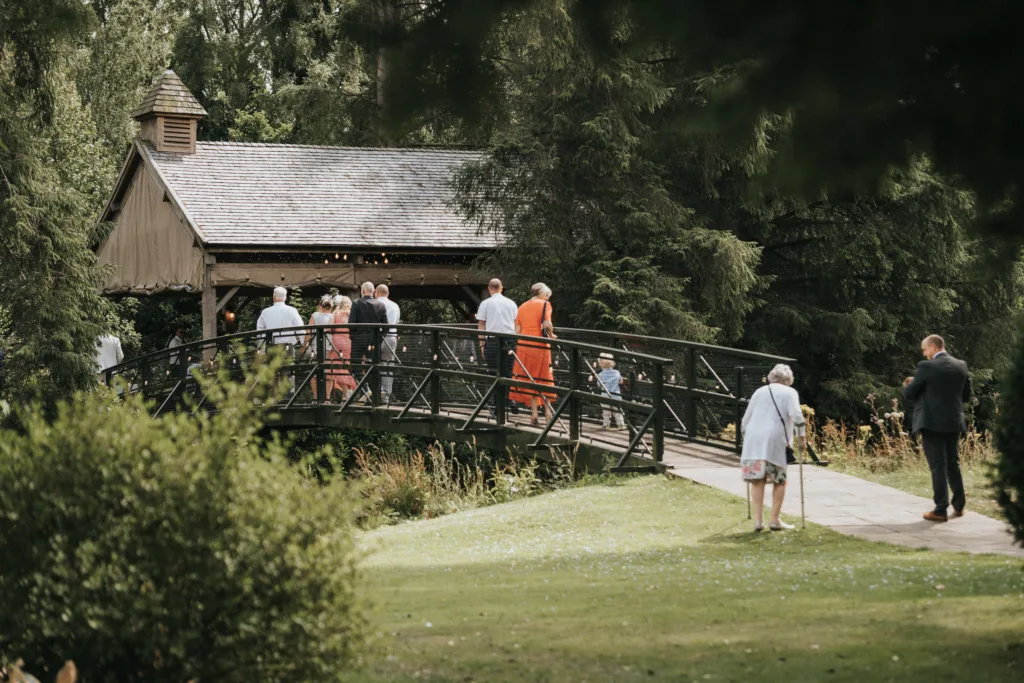 A group of people stand and walk on a curved footbridge at Hackness Grange near Scarborough, leading to a wooden pavilion surrounded by lush green trees. In the garden gathering, a wedding photographer captures moments as guests stroll along the path. © Aimee Lince Photography - Wedding photographer in Lincolnshire, Yorkshire & Nottinghamshire