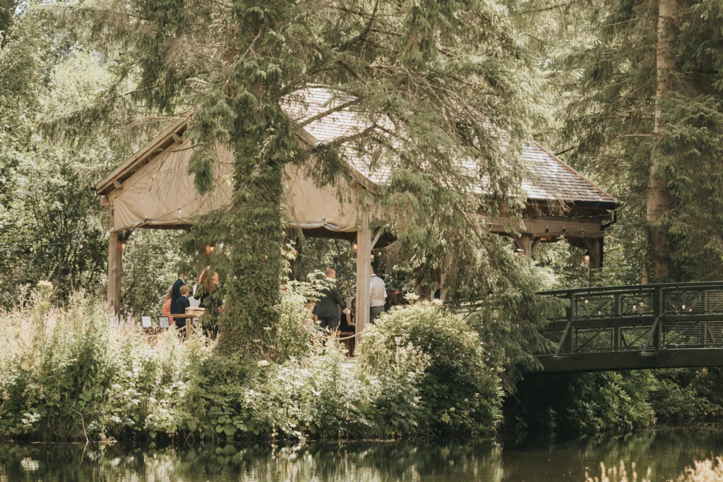 A group of people sit under a wooden gazebo at Hackness Grange, surrounded by greenery and tall pine trees. Next to the gazebo, a small arched bridge crosses a calm pond, while sunlight filters through—ideal for a wedding photographer in Scarborough. © Aimee Lince Photography - Wedding photographer in Lincolnshire, Yorkshire & Nottinghamshire