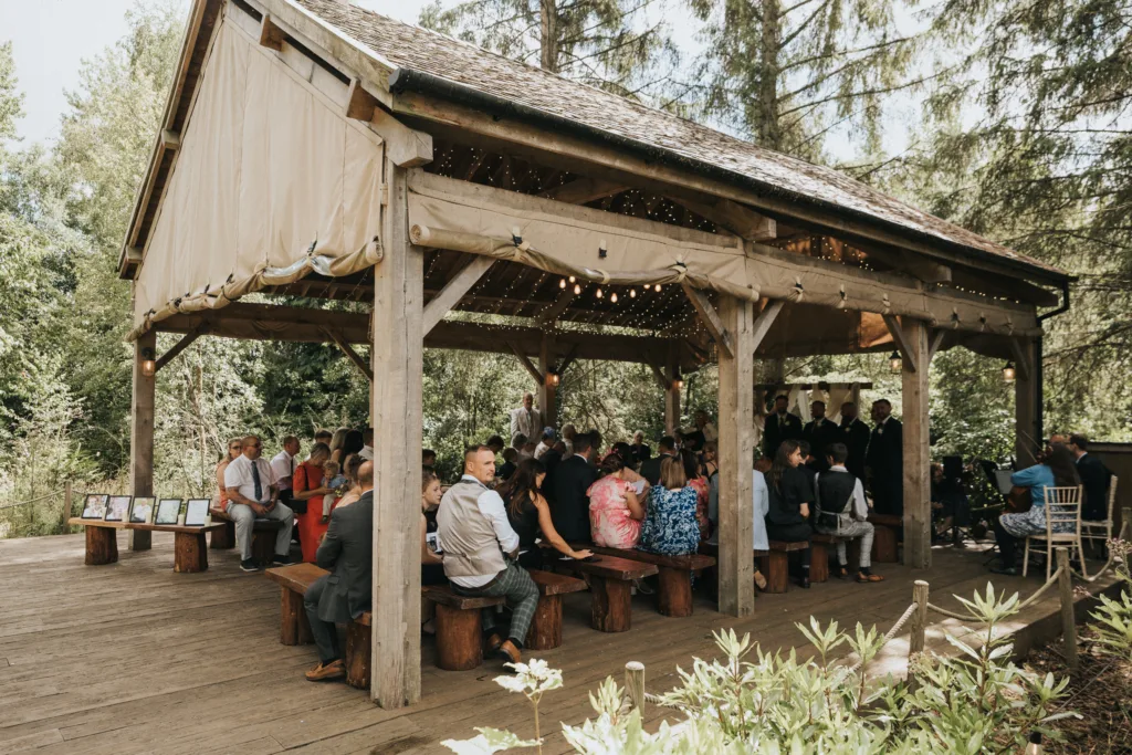 A group of people sit on wooden benches under a rustic wooden pavilion at Hackness Grange near Scarborough for an outdoor wedding ceremony, surrounded by trees. String lights twinkle above and framed photos welcome guests as sunlight filters through the greenery. © Aimee Lince Photography - Wedding photographer in Lincolnshire, Yorkshire & Nottinghamshire
