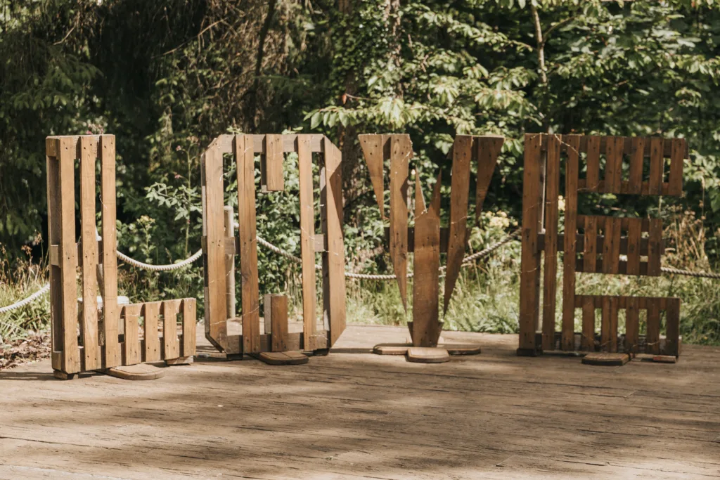 Large wooden letters spell "LOVE" on a wooden platform outdoors at Hackness Grange, near Scarborough. Sunlight filters through foliage, casting light and shadow—an ideal backdrop for any wedding photographer to capture a romantic moment. © Aimee Lince Photography - Wedding photographer in Lincolnshire, Yorkshire & Nottinghamshire