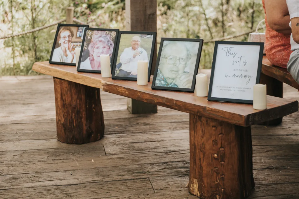A rustic wooden bench at Hackness Grange holds framed photos of elderly people and a sign reading, "This seat is reserved in memory." Four flameless candles sit between frames, creating a heartfelt scene outdoors with greenery—a perfect moment for any wedding photographer. © Aimee Lince Photography - Wedding photographer in Lincolnshire, Yorkshire & Nottinghamshire