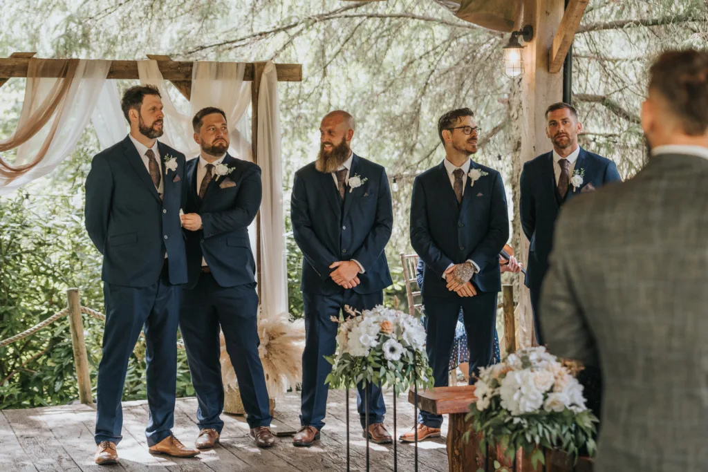 Five men in navy suits and brown shoes stand under a rustic wooden gazebo with draped fabric and flowers at Hackness Grange. Surrounded by greenery, these groomsmen await the outdoor ceremony, perfectly captured by a wedding photographer near Scarborough. © Aimee Lince Photography - Wedding photographer in Lincolnshire, Yorkshire & Nottinghamshire