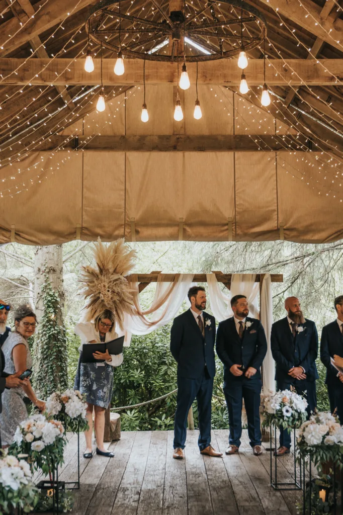 A rustic wooden wedding venue at Hackness Grange, near Scarborough, is decorated with string lights and white flowers. Two men in suits stand at the altar, with a wedding photographer capturing moments amid lush greenery and pampas grass. © Aimee Lince Photography - Wedding photographer in Lincolnshire, Yorkshire & Nottinghamshire