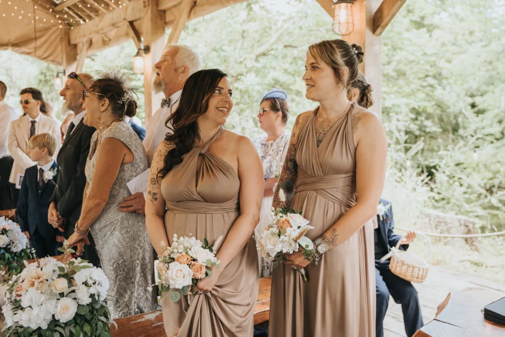 Two bridesmaids in taupe dresses hold bouquets of white and blush flowers, smiling at each other during a Hackness Grange wedding ceremony. Guests in formal attire gather under a wooden pavilion adorned with flowers and string lights near Scarborough. © Aimee Lince Photography - Wedding photographer in Lincolnshire, Yorkshire & Nottinghamshire