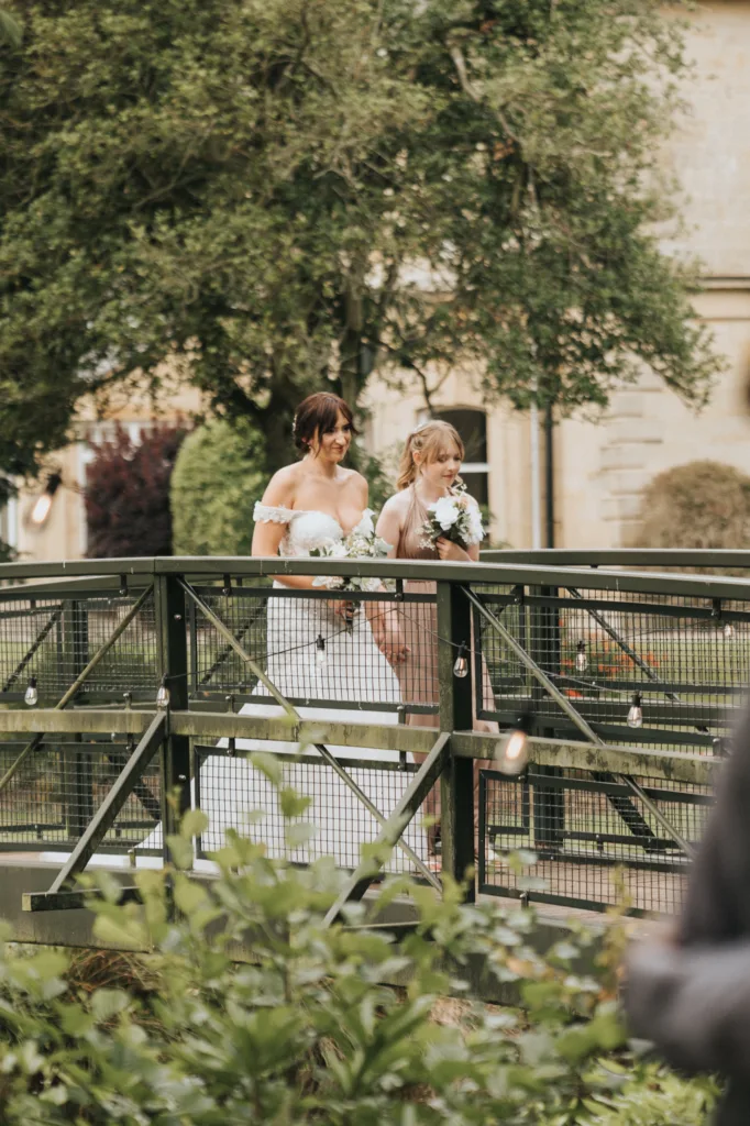 Two women, one in an off-shoulder white wedding gown and the other in taupe, walk side by side across a metal bridge at Hackness Grange. Captured by a Scarborough wedding photographer, they smile amid lush greenery and a stone building. © Aimee Lince Photography - Wedding photographer in Lincolnshire, Yorkshire & Nottinghamshire