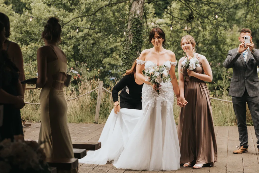 A bride in a white gown holds hands with a smiling bridesmaid in a taupe dress as another woman adjusts the bride’s train. Guests, including a man taking a photo, stand nearby at Hackness Grange near Scarborough, with green foliage behind. © Aimee Lince Photography - Wedding photographer in Lincolnshire, Yorkshire & Nottinghamshire