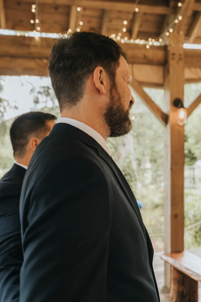 A man in a dark suit with a short beard stands in profile under a wooden pavilion decorated with string lights at Hackness Grange, near Scarborough. Another person in a suit stands beside him amid greenery—perfect for any wedding photographer. © Aimee Lince Photography - Wedding photographer in Lincolnshire, Yorkshire & Nottinghamshire