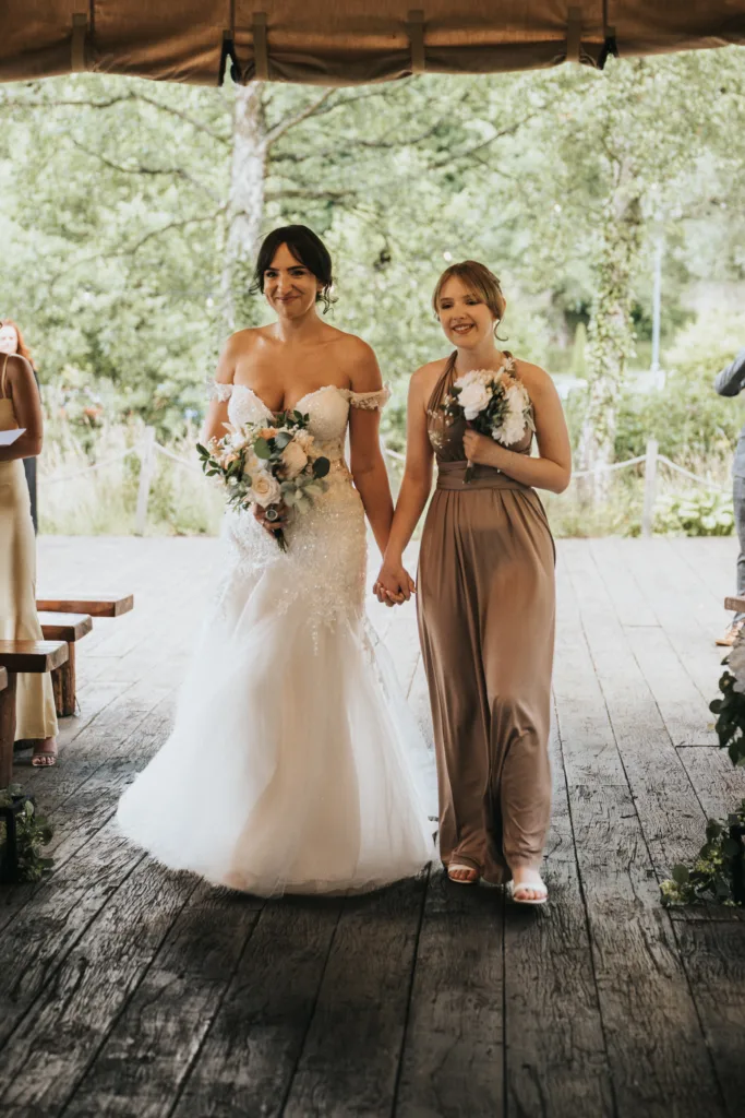 A bride in a white, off-shoulder gown walks hand-in-hand with a smiling bridesmaid in taupe at Hackness Grange, Scarborough. Captured by a wedding photographer, they stroll on a rustic wooden floor beneath a canopy with lush greenery behind them. © Aimee Lince Photography - Wedding photographer in Lincolnshire, Yorkshire & Nottinghamshire