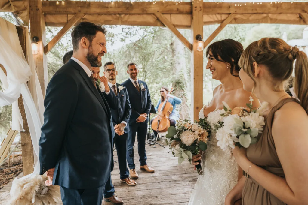 A bride in a white dress holds flowers and smiles at the groom under a wooden pavilion at Hackness Grange. Bridesmaids, groomsmen, and a wedding photographer capture the warmly lit scene, set amid lush greenery as a cellist plays in the background. © Aimee Lince Photography - Wedding photographer in Lincolnshire, Yorkshire & Nottinghamshire