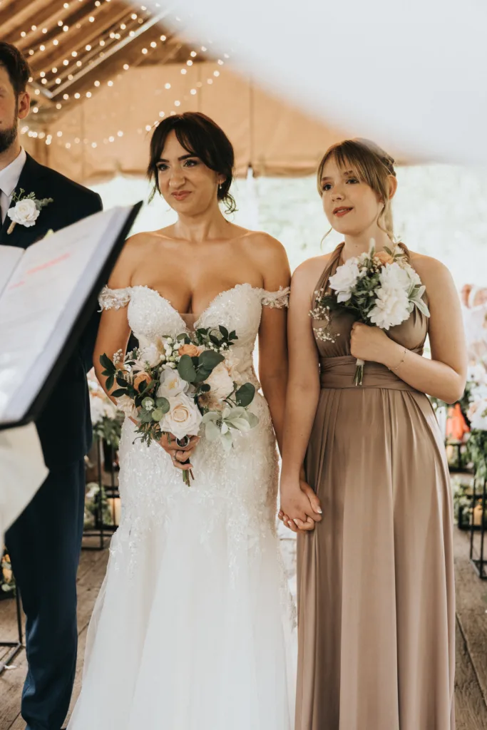 A bride in a white lace off-shoulder gown holds a bouquet and stands hand-in-hand with her bridesmaid beneath string lights at Hackness Grange, Scarborough—a perfect moment for any wedding photographer. Part of another suited guest and a music stand are visible at left. © Aimee Lince Photography - Wedding photographer in Lincolnshire, Yorkshire & Nottinghamshire