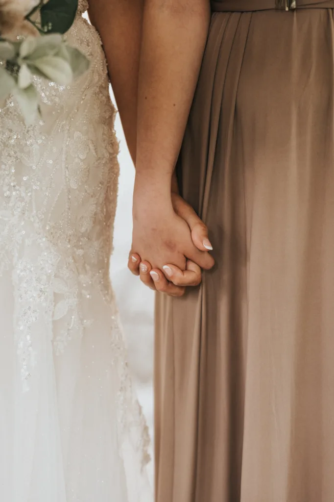 Two women stand side by side, holding hands. One wears a white, intricately beaded lace wedding dress, the other a taupe pleated gown. Captured by a wedding photographer at Hackness Grange near Scarborough, their closeness and support are beautifully evident. © Aimee Lince Photography - Wedding photographer in Lincolnshire, Yorkshire & Nottinghamshire