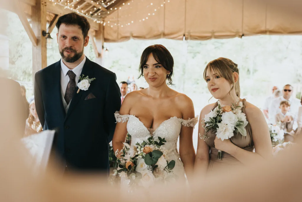 A bride in an off-shoulder white gown stands between a man in a dark suit and a bridesmaid in taupe, all holding bouquets. Indoors at Hackness Grange near Scarborough, under string lights—captured perfectly by the wedding photographer. © Aimee Lince Photography - Wedding photographer in Lincolnshire, Yorkshire & Nottinghamshire