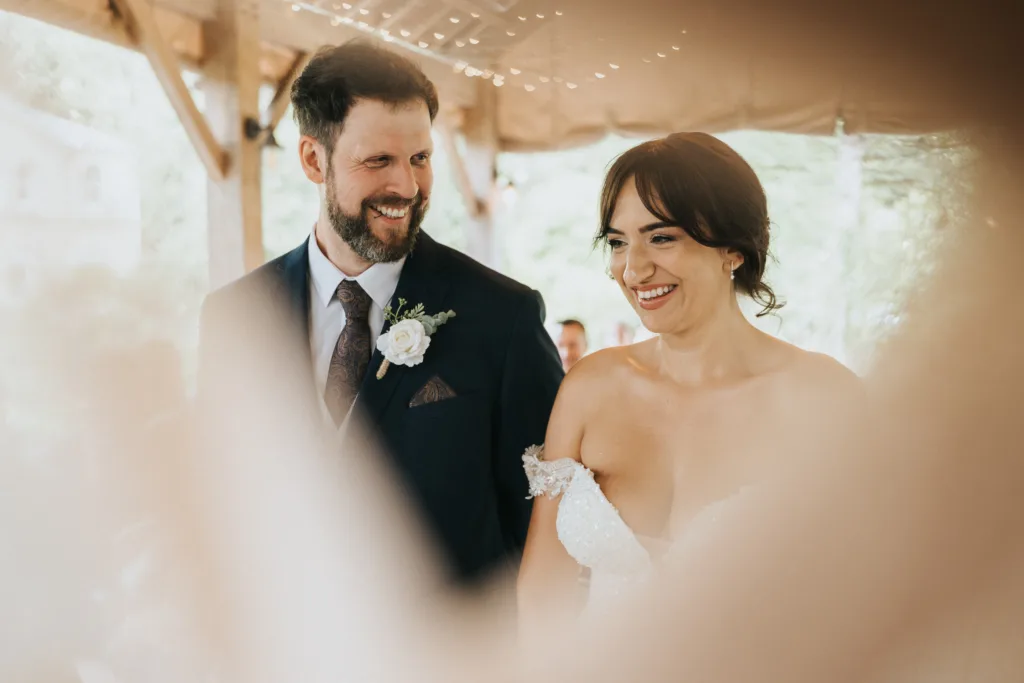 A bride and groom stand closely together, smiling at each other at Hackness Grange near Scarborough. The soft, blurred foreground frames them in a warm, candid moment captured perfectly by their wedding photographer. © Aimee Lince Photography - Wedding photographer in Lincolnshire, Yorkshire & Nottinghamshire