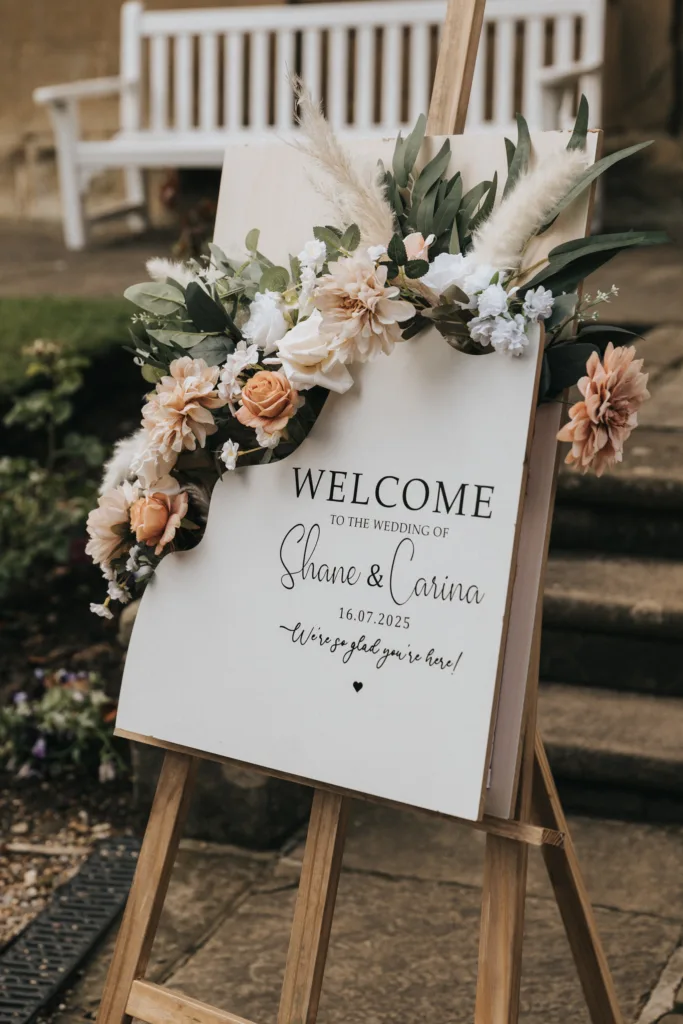 A wedding welcome sign on an easel reads, “Welcome to the wedding of Shane & Carina, 16.07.2025, we’re glad you’re here!” Decorated with cream and peach flowers at Hackness Grange near Scarborough—a perfect spot for any wedding photographer. © Aimee Lince Photography - Wedding photographer in Lincolnshire, Yorkshire & Nottinghamshire