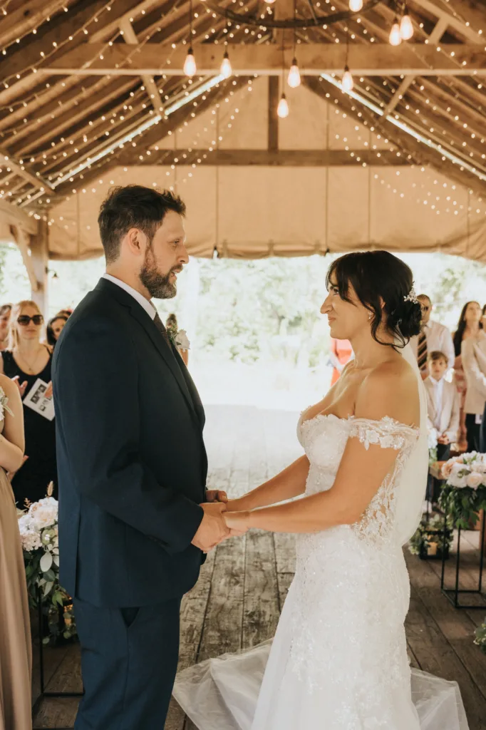A bride and groom stand facing each other, holding hands, during an outdoor wedding ceremony at Hackness Grange near Scarborough, under a wooden pavilion with string lights. A wedding photographer captures the moment as guests watch on. © Aimee Lince Photography - Wedding photographer in Lincolnshire, Yorkshire & Nottinghamshire
