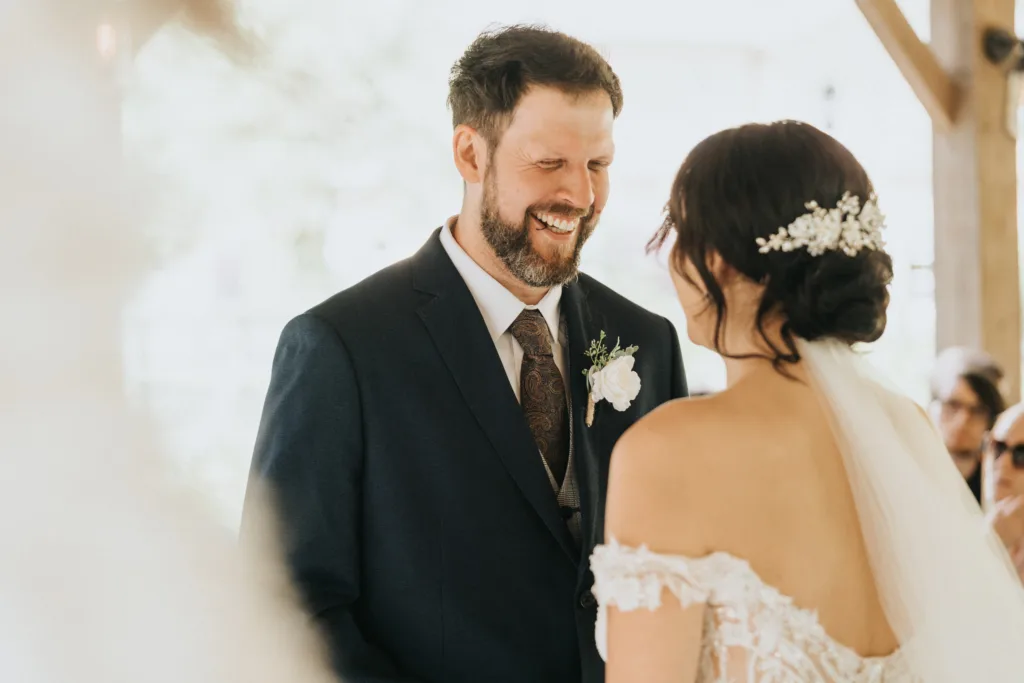 A wedding photographer captures a groom in a dark suit and patterned tie smiling at his bride, who wears an off-the-shoulder lace dress and floral veil. They stand facing each other at their Hackness Grange outdoor ceremony near Scarborough. © Aimee Lince Photography - Wedding photographer in Lincolnshire, Yorkshire & Nottinghamshire