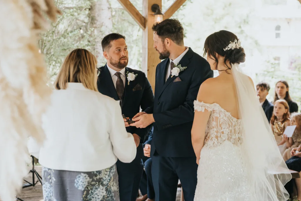A bride in a lace off-shoulder gown and veil stands beside a groom in a navy suit, holding hands during an outdoor Hackness Grange ceremony near Scarborough, as a wedding photographer captures the moment while guests watch attentively. The scene is bright and natural. © Aimee Lince Photography - Wedding photographer in Lincolnshire, Yorkshire & Nottinghamshire