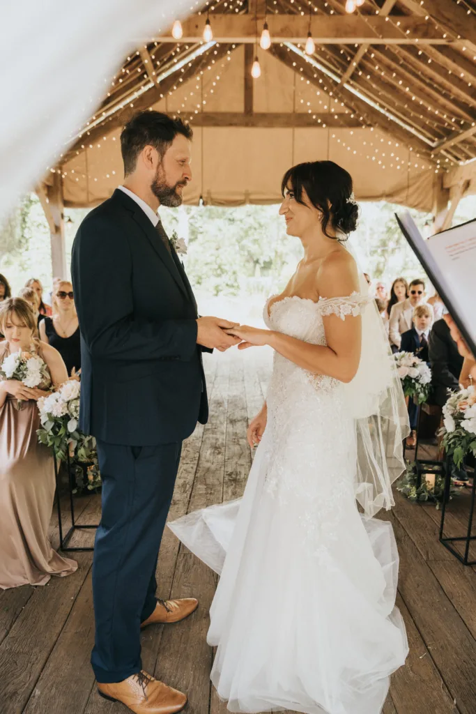A bride in a white off-shoulder lace gown and a groom in a navy suit exchange vows under a wooden pavilion at Hackness Grange. Guests watch the ceremony surrounded by greenery, with a Scarborough wedding photographer capturing every moment. © Aimee Lince Photography - Wedding photographer in Lincolnshire, Yorkshire & Nottinghamshire