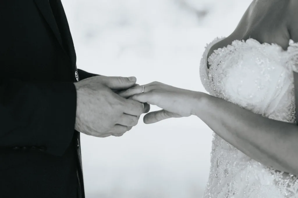 A close-up, black-and-white photo by a Hackness Grange wedding photographer. The groom, in a dark suit, places a ring on the bride’s finger. She wears a lace-embroidered dress. Only their torsos and hands are visible; the background is blurred. © Aimee Lince Photography - Wedding photographer in Lincolnshire, Yorkshire & Nottinghamshire