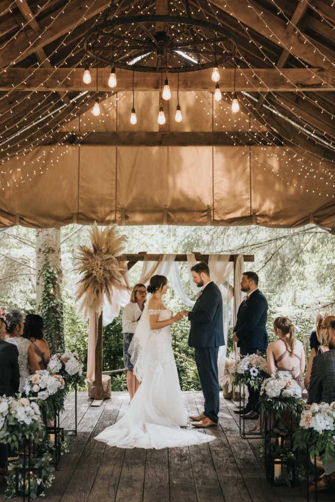 A bride and groom stand facing each other under a rustic wooden gazebo at Hackness Grange, adorned with hanging lights and floral arrangements. Guests look on as a wedding photographer captures the vows, with pampas grass decorating the altar behind them. © Aimee Lince Photography - Wedding photographer in Lincolnshire, Yorkshire & Nottinghamshire