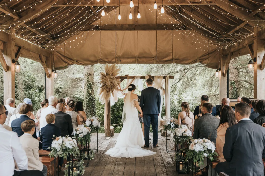 A bride and groom stand at an outdoor wooden altar under a decorated pavilion at Hackness Grange near Scarborough, surrounded by guests. White flowers, greenery, and string lights set the scene for a warm, rustic wedding—perfect for any wedding photographer. © Aimee Lince Photography - Wedding photographer in Lincolnshire, Yorkshire & Nottinghamshire