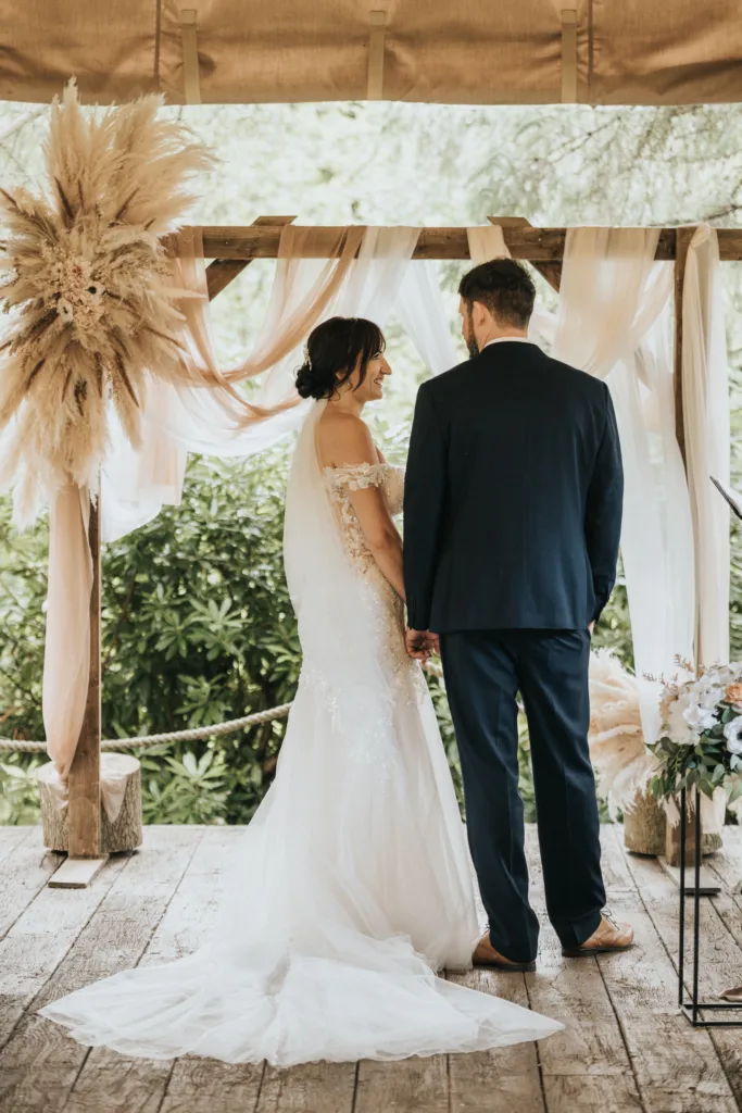 A bride in a white lace gown and veil stands with her groom in a navy suit at Hackness Grange. Sunlight filters through greenery as their wedding photographer captures the moment beneath a pampas-adorned arch on the wooden platform near Scarborough. © Aimee Lince Photography - Wedding photographer in Lincolnshire, Yorkshire & Nottinghamshire