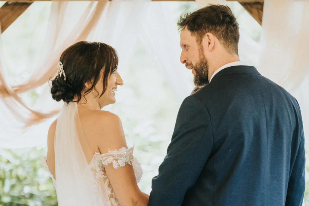 A bride in a white dress with lace and veil smiles at her groom in a navy suit under the softly lit wedding arch at Hackness Grange. A Scarborough wedding photographer captures their joyful moment, hands clasped amid lush greenery. © Aimee Lince Photography - Wedding photographer in Lincolnshire, Yorkshire & Nottinghamshire