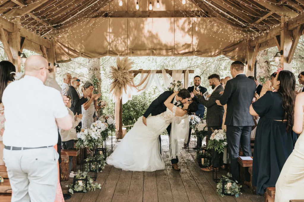 A bride and groom share a kiss at the altar during an outdoor rustic wedding ceremony at Hackness Grange, surrounded by cheering guests. The wooden pavilion is decorated with draped fabric, hanging lights, flowers, and greenery. © Aimee Lince Photography - Wedding photographer in Lincolnshire, Yorkshire & Nottinghamshire