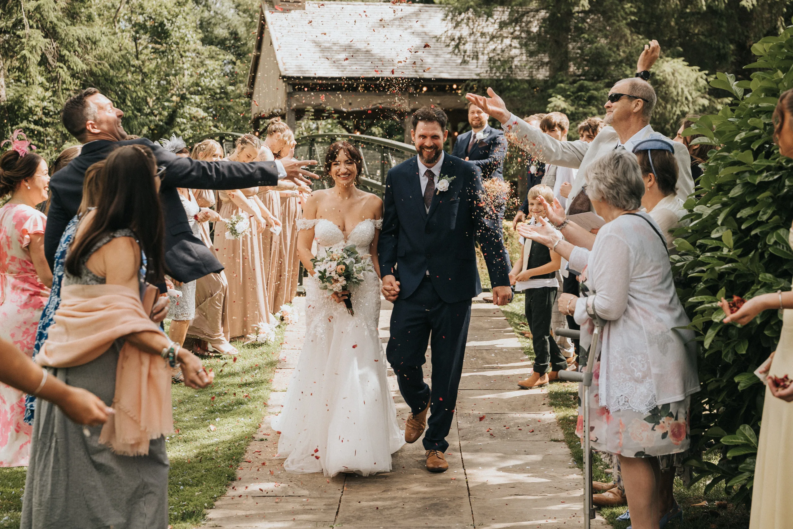 shane-carina-wedding-photography-scarborough-hackness-grange-photographer-copyright-aimee-lince-270-of-649-©-aimee-lince-photography-16-07-2025 A bride and groom, smiling and holding hands, walk down an outdoor aisle at Hackness Grange as guests toss flower petals in celebration. Captured by a Scarborough wedding photographer, the festive scene features greenery and a rustic building in the background. © Aimee Lince Photography - Wedding photographer in Lincolnshire, Yorkshire & Nottinghamshire