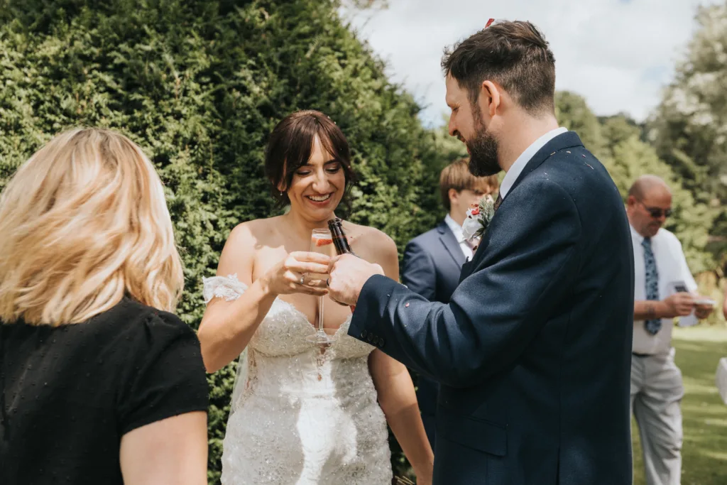 A bride in a lace wedding dress and a groom in a navy suit laugh together outdoors at Hackness Grange near Scarborough, clinking drink bottles. The joyful, candid moment is perfectly captured by their wedding photographer as guests celebrate in the sunlight. © Aimee Lince Photography - Wedding photographer in Lincolnshire, Yorkshire & Nottinghamshire