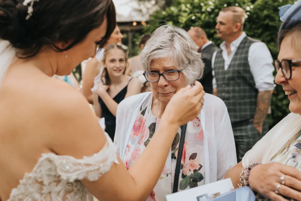 A bride in a white dress gently wipes tears from an elderly woman's face at Hackness Grange. The older woman wears glasses, a white cardigan, and a floral scarf. Nearby, smiling guests stand outdoors amid greenery—captured by a wedding photographer. © Aimee Lince Photography - Wedding photographer in Lincolnshire, Yorkshire & Nottinghamshire