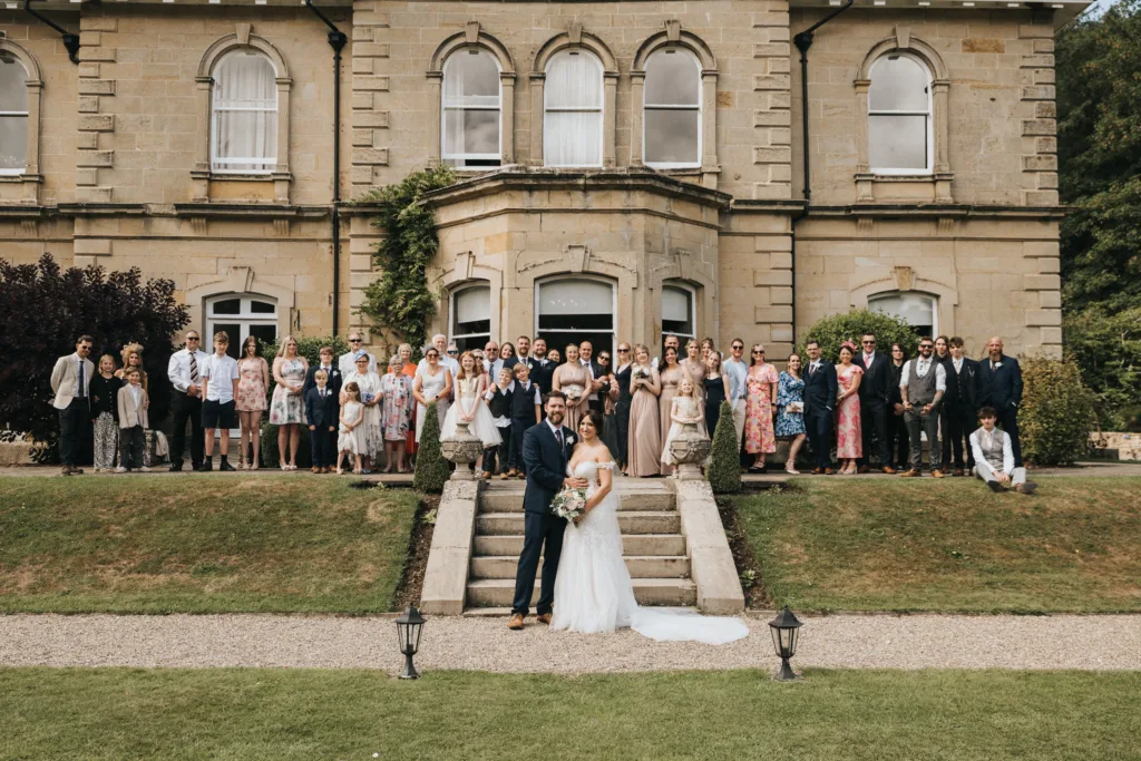 A bride and groom stand on steps in front of the grand Hackness Grange near Scarborough, holding hands and smiling at the camera. Their wedding photographer captures guests posing behind them on tiered grass lawns, dressed in elegant attire. © Aimee Lince Photography - Wedding photographer in Lincolnshire, Yorkshire & Nottinghamshire